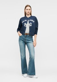 Γυναίκα που φορεί μπλε σκούρο cardigan Ralph Lauren με λογότυπο, ανοιχτό γκρι πουκάμισο, καφέ ζώνη, τζιν παντελόνι με φαρδιά μπατζάκια και λευκά αθλητικά παπούτσια, στέκεται με το ένα χέρι στην τσέπη.