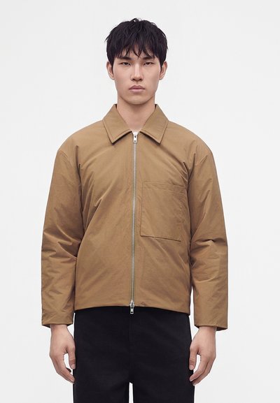 Jeune homme portant une veste zippée marron clair avec une poche poitrine et un pantalon noir, debout devant un fond blanc uni.