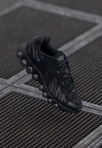 Sapatilha atlética preta com um design ondulado, superfície texturizada, fecho de atacadores na parte da frente e uma sola almofadada proeminente com seções circulares.