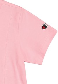 Camiseta de algodón rosa claro con mangas cortas, que presenta un parche de logo en rojo y negro en la manga izquierda. Detalles de costura limpia visibles.