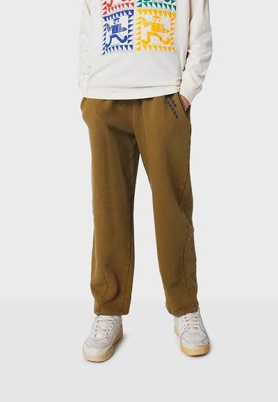 Braune Baumwoll-Sweatpants mit Seitentaschen, elastischem Bund und blauem Stickdetail. Getragen mit einem hellen grafischen Sweatshirt und weißen Turnschuhen.