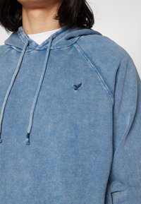 Ljusblå hoodie i mjukt, strukturert tyg med en känguruficka. Har en liten marinblå fåglogotyp på bröstet och justerbara dragsnören.