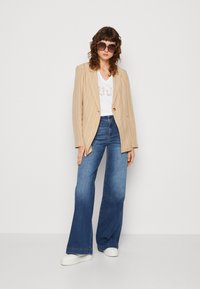 Blazer a righe beige sopra una t-shirt bianca con grafica, abbinato a jeans blue a zampa e sneakers bianche. Grandi occhiali da sole quadrati completano l'outfit.