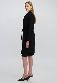 Cappotto nero con vita cintrata, colletto a revers e due tasche. Tessuto liscio, design fino al ginocchio. Indossato sopra un vestito nero e tacchi.