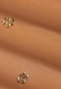 Tissu de couleur beige chaud présentant une décoration florale dorée détaillée avec des sequins, placée sur un fond lisse et mat.