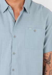 Chemise bleu clair à manches courtes avec boutons, fabriquée en tissu texturé, dotée d'une poche poitrine et d'une étiquette. Les boutons sont ronds et contrastés.