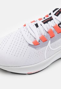 Gros plan sur une chaussure de course Nike blanche avec des passants de lacets corail, des lacets blancs, une tige en mesh respirant et un détail de semelle « Air Zoom ».