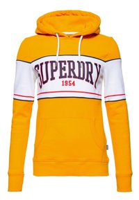 Sudadera amarilla con capucha y una franja blanca y negra. En la parte frontal presenta "SUPERDRY" y "1954" en letras rojas. Tiene un bolsillo canguro. Mangas largas.