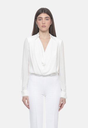 Donna con lunghi capelli scuri che indossa una camicetta bianca drappeggiata con colletto e pantaloni bianchi a vita alta, in piedi davanti a uno sfondo semplice.