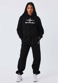 Zwarte zip-up hoodie met een wit logo, gecombineerd met zwarte sweatpants. Beide gemaakt van zachte stof, met een relaxte pasvorm en voorzakken.