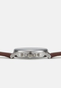 Montre en acier inoxydable avec un boîtier rond, un bracelet en cuir marron, une couronne ronde et deux poussoirs sur le côté. Design minimaliste avec une finition lisse.