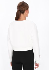 Sudadera corta blanca con mangas largas, puños acanalados y cuello redondo. Presenta un logo en la manga izquierda.