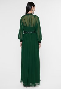 Robe en chiffon vert longue avec des manches transparentes, une taille perlée et une jupe fluide. Le dos présente un col haut et un plissage subtil.