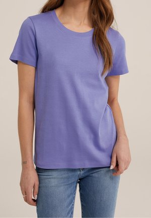 Kobieta ubrana w prosty lawendowy t-shirt z krótkim rękawem i niebieskie dżinsy, stoi z jedną ręką luźno opuszczoną, a drugą trzymającą rąbek koszulki.