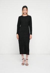 Vestido preto de manga longa com um padrão de riscas texturizadas. Comprimento até à metade da panturrilha, com um decote redondo e um corte ajustado. Sandálias pretas de salto.