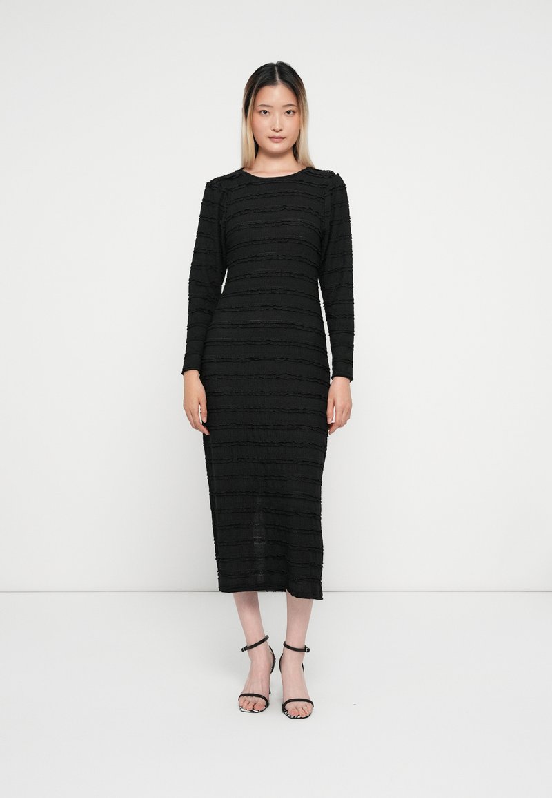 Vestido preto de manga longa com um padrão de riscas texturizadas. Comprimento até à metade da panturrilha, com um decote redondo e um corte ajustado. Sandálias pretas de salto.