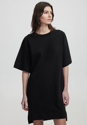 Schwarzes, oversized T-Shirt-Kleid mit kurzen Ärmeln, rundem Halsausschnitt, seitlichen Schlitzen und glatter Textur. Einfaches Design ohne Muster oder Verzierungen.