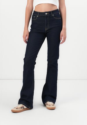 Jean flare bleu foncé à taille haute, avec poches avant et un détail en bouton doré. Le tissu a une texture lisse avec des surpiqûres contrastées.