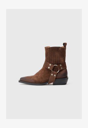Braunes Wildleder-Ankle-Boot mit spitzer Zehenform, dekorativer Naht und einem Riemen mit einem Metallring und silbernen Nieten. Niedriger Absatz.