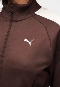 Καφέ τζάκετ με φερμουάρ και ψηλό γιακά, υφασμάτινη υφή, λευκή λεπτομέρεια στον ώμο και λευκό λογότυπο της Puma στο αριστερό στήθος.