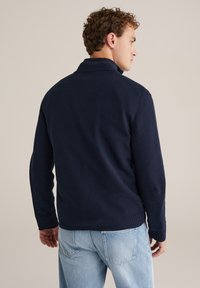 Teksturirani mornarsko plavi pamučni pulover s visokim ovratnikom i rebrastim manžetnama, prikazan s leđa, u kombinaciji sa svijetloplavim trapericama.