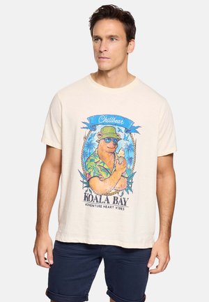 Hombre con camiseta beige claro con un oso de dibujos animados colorido, con gafas de sol y sombrero, sosteniendo un helado, combinado con pantalones cortos azul oscuro.