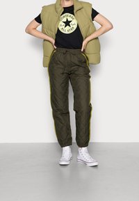 Olijfgroene gewatteerde vest over een zwart T-shirt met een gouden logo, gecombineerd met donkergroene gewatteerde broek met gele zijkanten en witte sneakers.