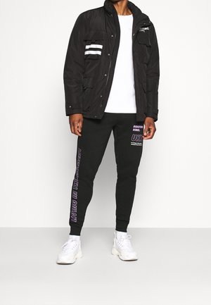Sort vandtæt jakke med to frontlommer og hvide stribede detaljer, parret med sorte sweatpants med lilla tekst og hvide sneakers.
