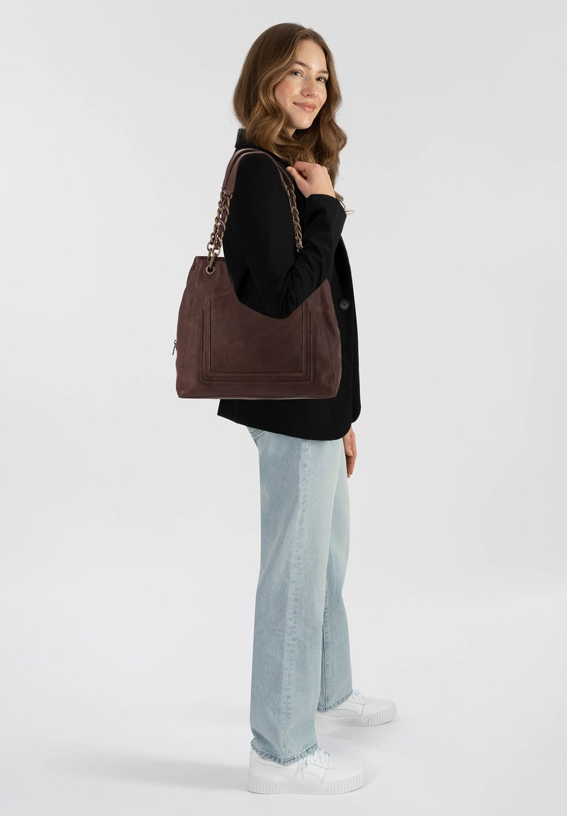 Donna con blazer nero e jeans chiari in piedi di lato, tiene una grande borsa in pelle marrone con catene sulle spalle.