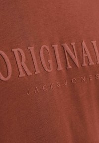 Rudai spalvos medvilnės marškinėliai su iškilniuoju užrašu "ORIGINAL" ir "JACK & JONES". Lygus audinys, trumpomis rankovėmis ir apvalia iškirpte.