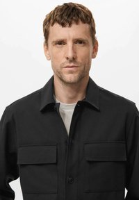 Moški s kratkimi rjavimi lasmi, oblečen v črno srajco na gumbe z dvema prsima žepoma, oblečeno čez belo majico, obrne se proti spredaj na belem ozadju.