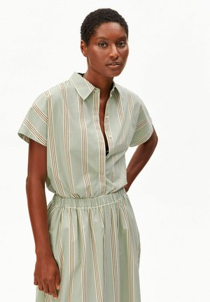 Vrouw met kort krullend haar, gekleed in een lichtgroene jurk met verticale bruine en witte strepen, staand tegen een witte achtergrond.