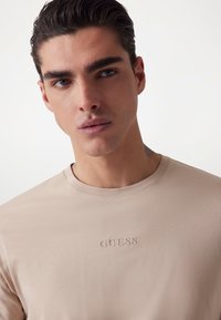 Light beige cotton t-shirt featuring a crew neckline and embossed "GUESS" logo on the chest. Smooth texture with short sleeves.