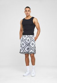 Black ribbed tank top paired with patterned white shorts featuring black accents. Model wears white sneakers and white socks, hands in pockets.