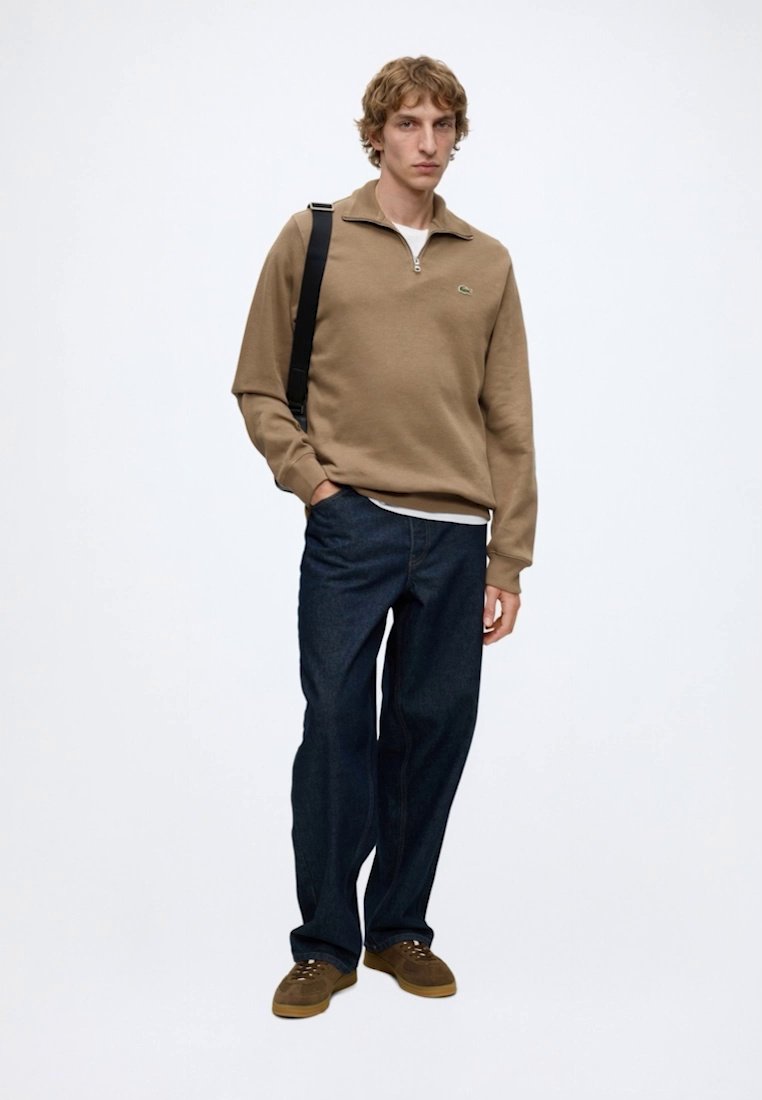 Jeune homme debout portant un pull beige à fermeture éclair quart, un jean bleu foncé, des chaussures marron et un sac bandoulière noir sur fond blanc.
