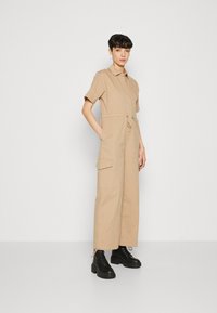 Beige utility jumpsuit with short sleeves, front zipper, and drawstring waist. Features side pockets and tapered legs. Paired with black boots.