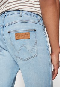 Lichtblauwe denim jeans hebben een leren Wrangler-label op de achterzak, met contrasterende stiksels en een gegolfd zakontwerp.