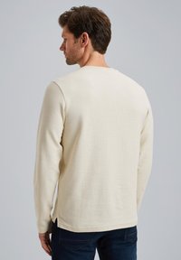 Helle beige Langarmshirt mit strukturiertem Strickdesign, Rundhalsausschnitt und seitlichen Schlitzen, kombiniert mit dunkelblauen Jeans.