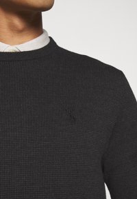 Suéter gris oscuro con textura y cuello redondo, con un sutil logo negro bordado en el frente. Combinado con una camisa blanca con cuello debajo.