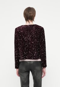 Asmuo su trumpais plaukais, vilkintis tamsiai purpurinį pailgintą švarką su blizgučiais ir juodas odines kelnes, stovi atsisukęs nugara prieš paprastą foną.