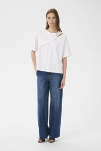 Balti marškinėliai su kampuotais iškirpimo elementais ir trumpomis rankovėmis, derinami su aukštos liemens, plačiomis mėlynomis džinsinėmis kelnėmis. Matoma tekstūruota džinsinio audinio faktūra.