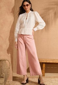 Blouse peplum blanche texturée avec des accents en dentelle, associée à un pantalon large rose. Le mannequin porte des lunettes de soleil surdimensionnées et des sandales à talons épais.