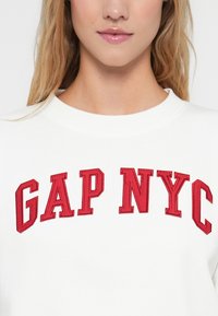 Biała bluza z czerwoną haftowaną napisem "GAP NYC" w wyrazistej, uczelnianej czcionce; okrągły dekolt i miękka faktura materiału.
