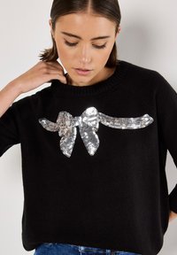 Maglione nero con un collo a costine e un dettaglio centrale a fiocco di paillettes. Il tessuto appare morbido e strutturato, accompagnato da una vestibilità comoda.