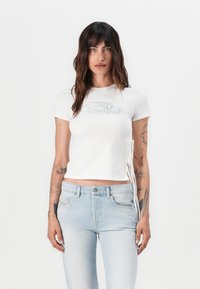 White cropped t-shirt with a round neckline and short sleeves. Features side ties and a faint graphic logo on the chest. Textured cotton fabric.