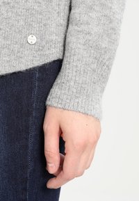 Grauer Strickpullover mit einem gerippten Muster, versehen mit einem kleinen silbernen Logoakzent und einem leicht schrägen Saum, kombiniert mit dunklem Denimstoff.