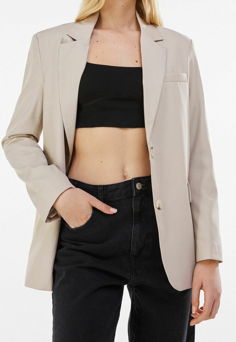 Femme portant un blazer beige, un top court noir et un jean noir taille haute, avec une main dans la poche du jean, sur fond blanc.