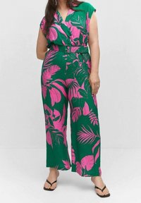 Donna che indossa una tuta verde con motivi di foglie tropicali rosa, vita cintrata e sandali neri infradito, in piedi davanti a uno sfondo neutro.