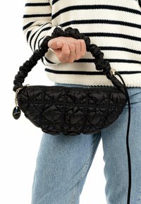 Borsa a tracolla nera trapuntata con una superficie testurizzata, dotata di un manico arricciato e decorata con un piccolo charm, tenuta da una persona con un maglione a righe.
