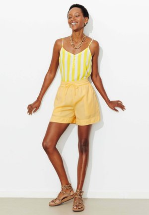 Femme souriante appuyée contre un mur blanc, portant un débardeur jaune rayé, un short jaune, un collier chaîne en or et des sandales imprimé léopard.